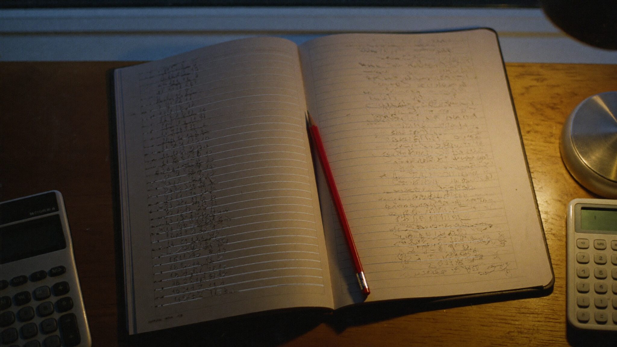Open ledger under a desk lamp with a red pencil resting across a column of revised figures