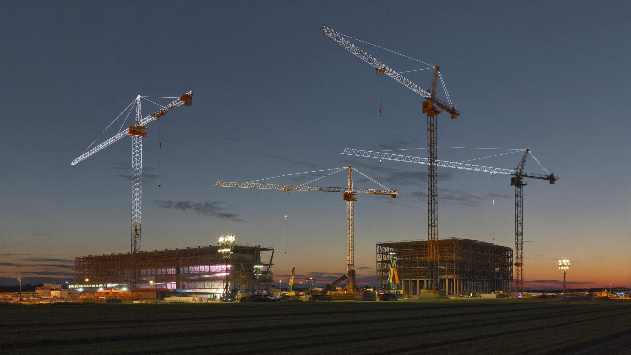 Deserted data-center construction site at dusk with four cranes standing over the pad under work lights
